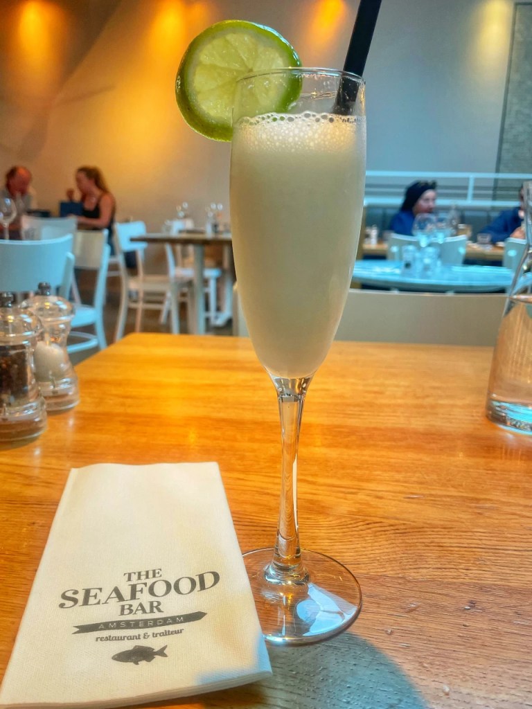 Et cocktailglass med en lys drink og en lime skive som dekorasjon, plassert på et trebord, med en meny fra The Seafood Bar i bakgrunnen.