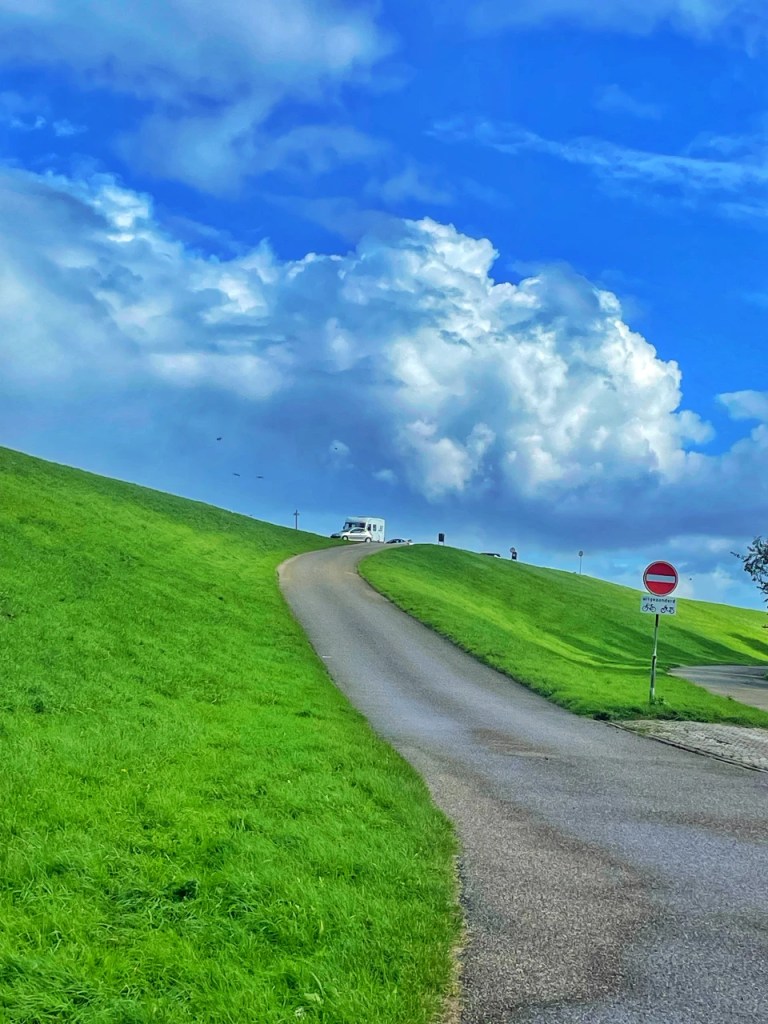 En vei som svinger oppover mot en grønn høyde, med en blå himmel og store, fluffy skyer. Det er et skilt for forbudt adgang og et skilt for sykkelstier.