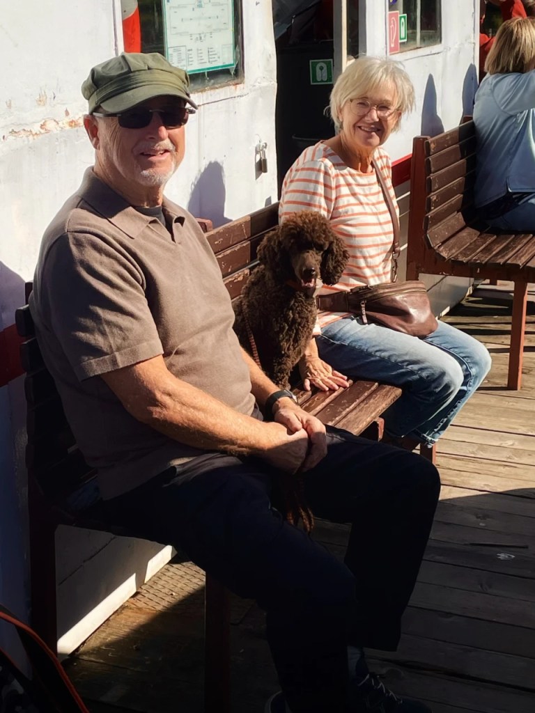 To personer og en hund sitter på en benk. En mann med briller og caps smiler mot kameraet, mens en kvinne i stripete genser smiler ved siden av ham. Hunden, en brun puddel, sitter mellom dem.