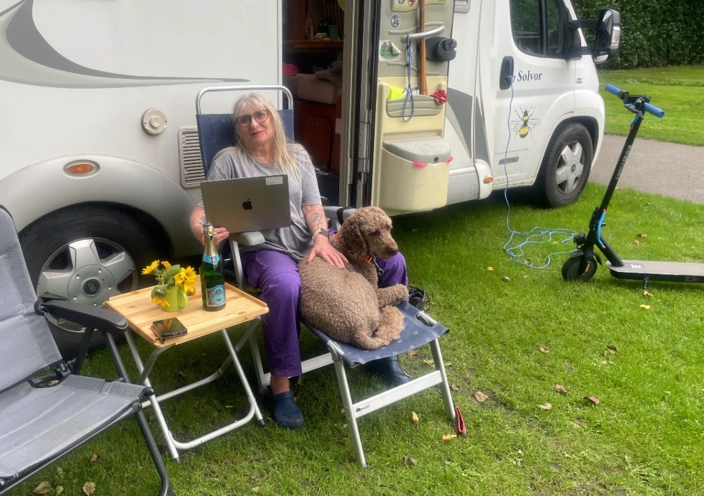 En kvinne sitter med en laptop på fanget, med en hund ved siden av seg, foran en campingvogn. Det er en liten blomsterpotte og en flaske med vann på et bord. Det er også en elektrisk sparkesykkel i bakgrunnen.
