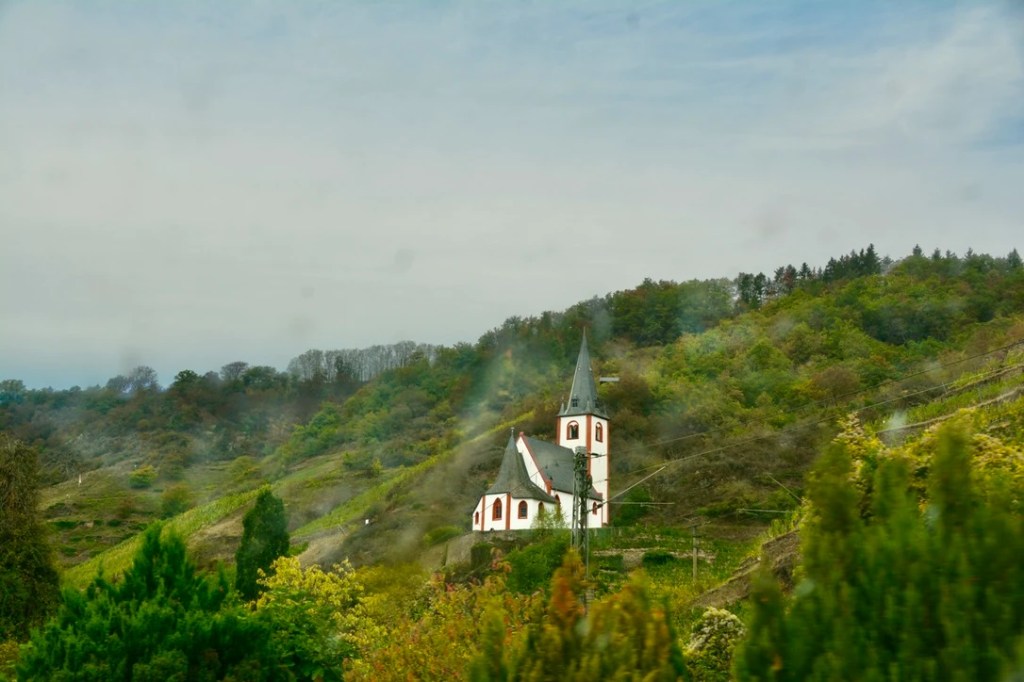 En liten kirke med spisstårn ligger på en skråning omgitt av grønne åser og trær.