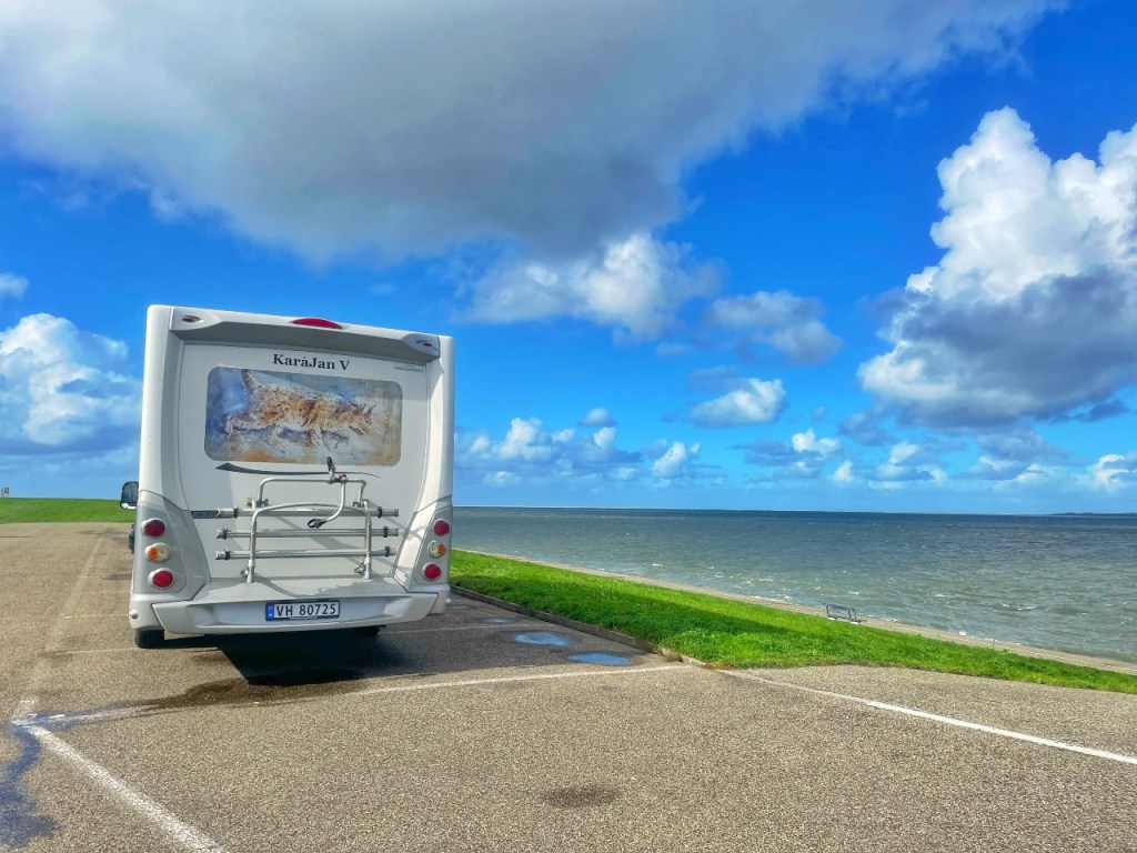 En campingvogn parkert ved vannkanten med en klar, blå himmel og hvite skyer i bakgrunnen.