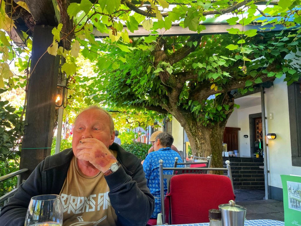 En eldre mann sitter ved et bord under et tre med grønne blader, mens han holder en gaffel i hånden. I bakgrunnen ses en annen person med blå skjorte. Det er en uteservering med solrike omgivelser.