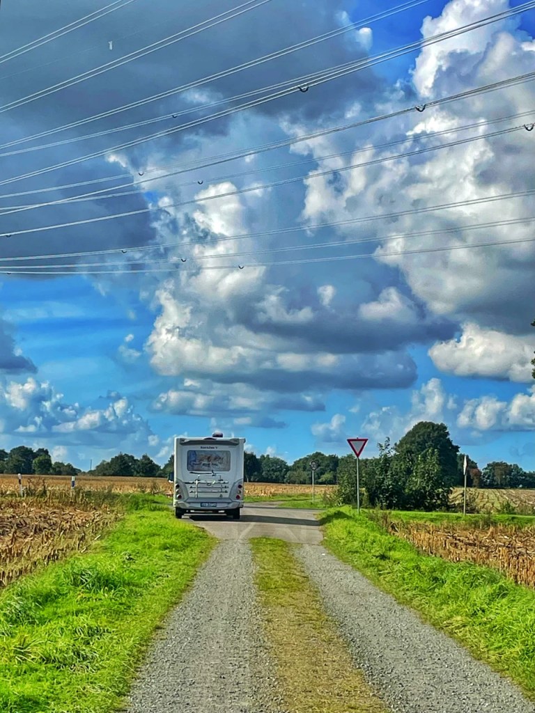 En campingvogn på en grusvei som strekker seg gjennom landskapet, med høye skyer og strømledninger over hodet.