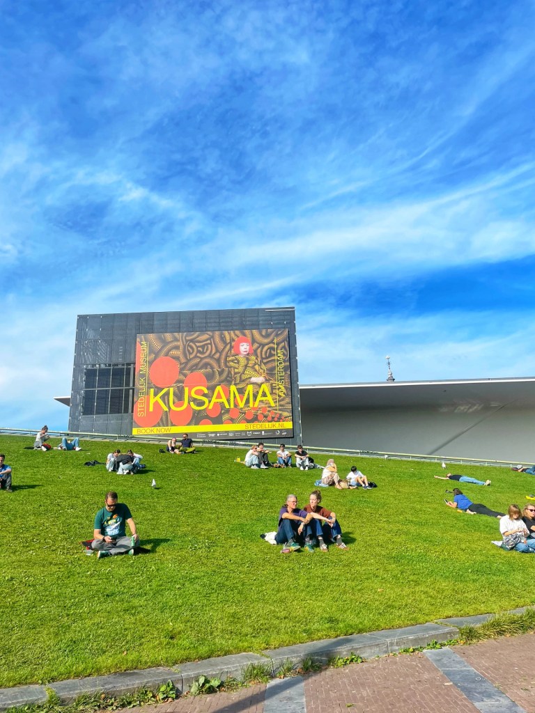 Folk sitter på gresset foran et stort KUSAMA-plakat ved et moderne bygg, med klar blå himmel i bakgrunnen.