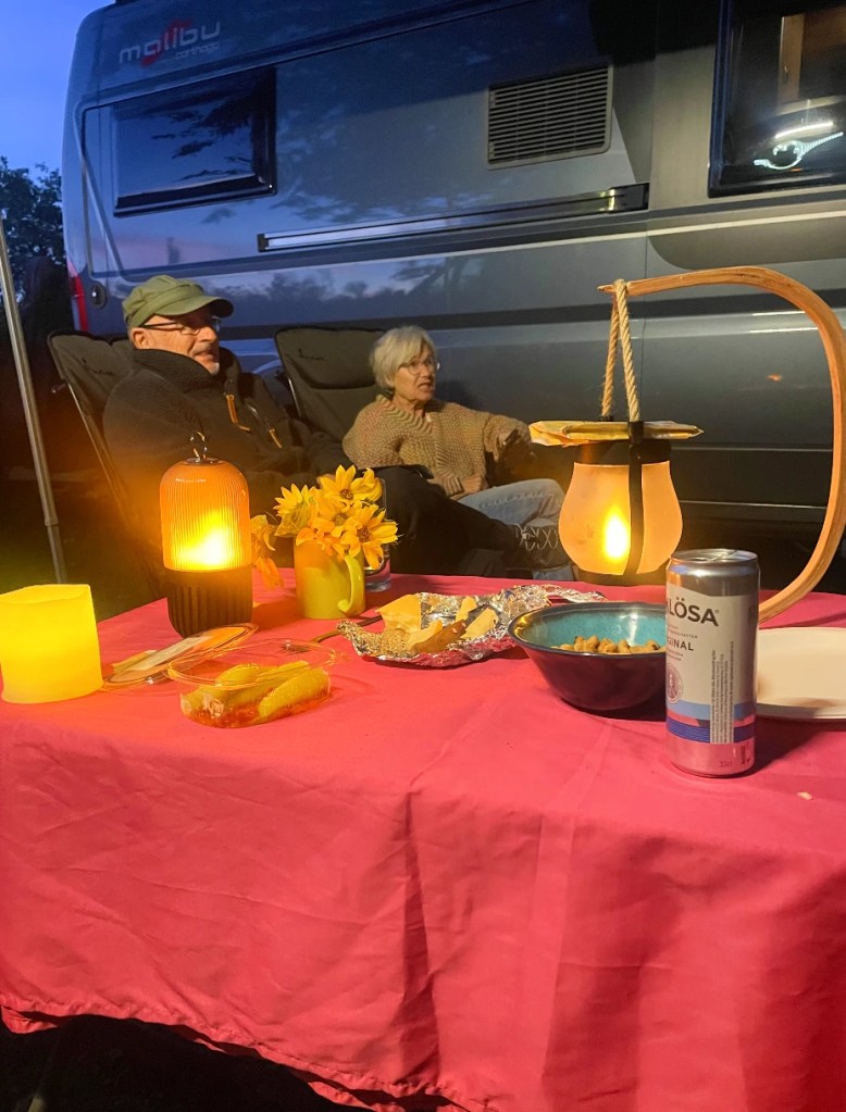 To personer sitter avslappet ved et bord med blomster og mat, om kvelden foran en camper.