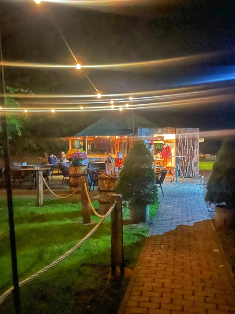Uteområde ved en restaurant om natten med dekorative lys og blomsterpotter, omgitt av busker og hagestoler.