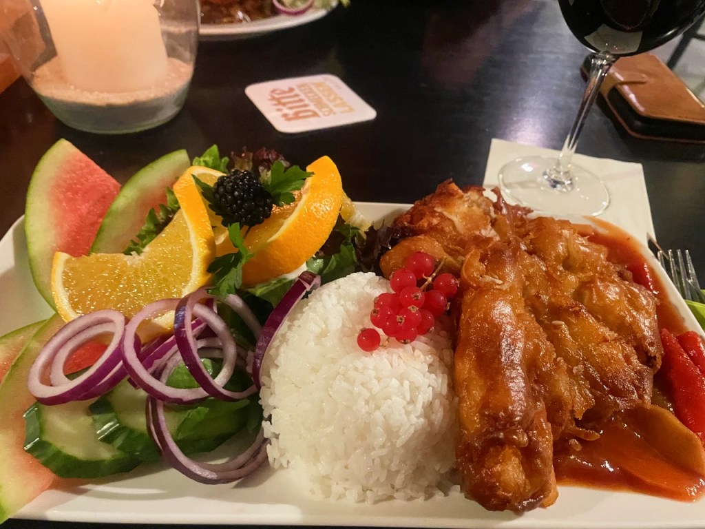En tallerken med stekt fisk, hvit ris, og friske grønnsaker, inkludert melonskiver, appelsinbiter, og rødløk. I bakgrunnen en vinrød glass og et stearinlys.