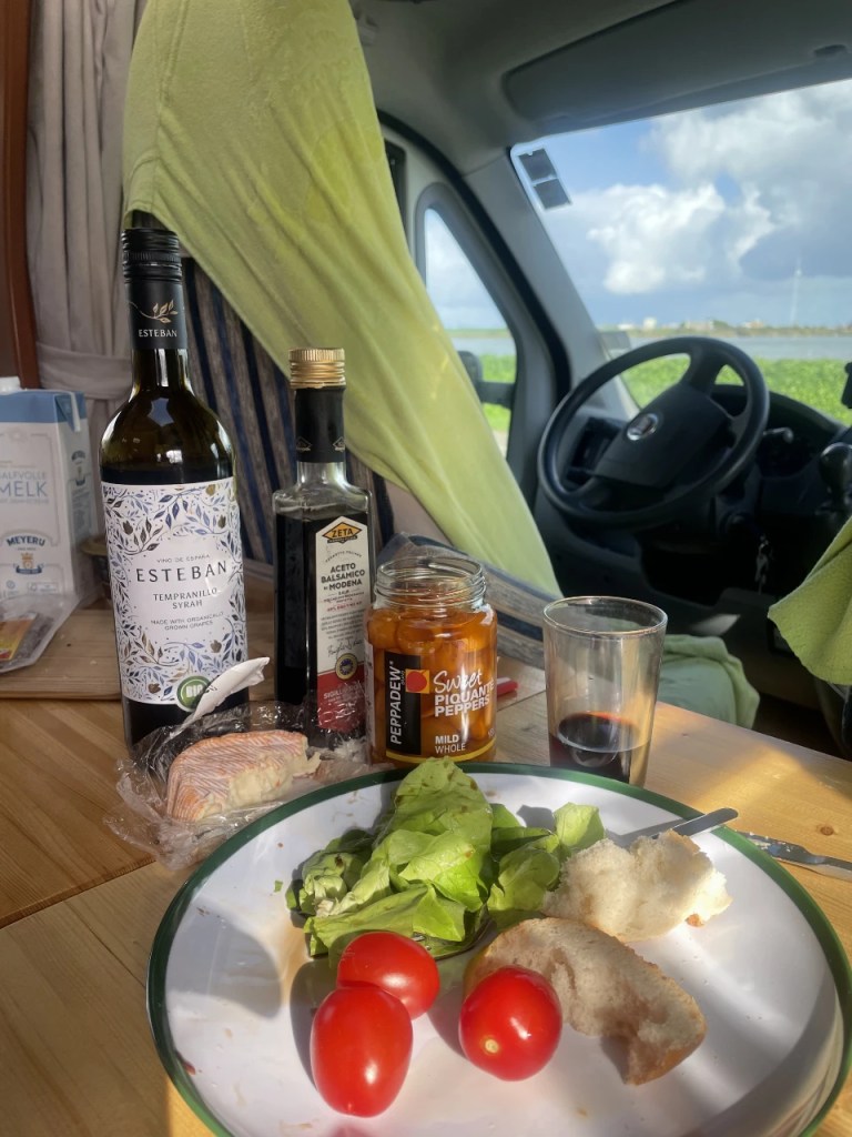 Et bord i en campingbil med en tallerken som inneholder tomater, salat og brød, sammen med flasker med vin og balsamico. Utsikt mot naturen utenfor vinduet.
