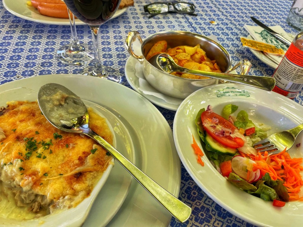 En tallerken med gratinert fiskeforus med potetmos, pyntet med persille, servert med en side av poteter og salat med tomat, agurk og gulrot. Et glass rødvin og en brusflaske er synlig i bakgrunnen.