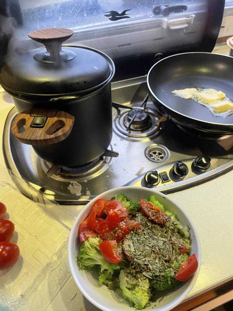 Kjøkkeninnstilling med en gryte som damper, en stekepanne med smør, og en bolle med salat bestående av brokkoli, tomater og krydder.