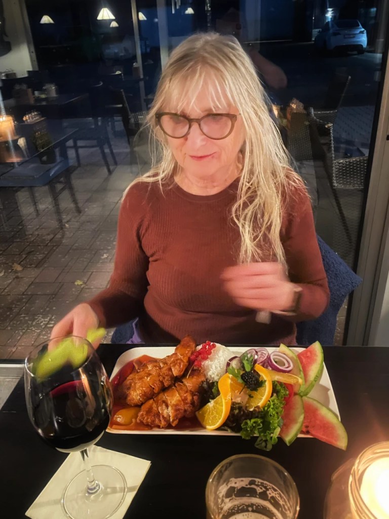 En kvinne sitter ved et bord med et måltid som inkluderer grillet kylling, ris, frukt og grønnsaker, mens hun holder et glass rødvin.