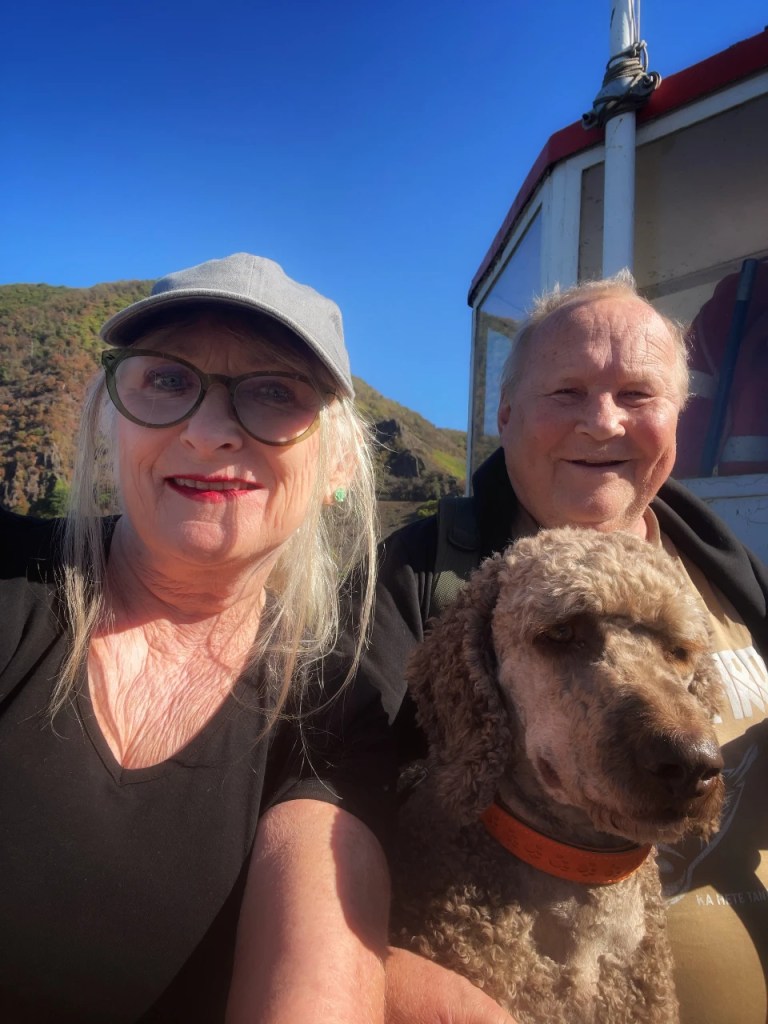 Selvportrett av en kvinne og en mann med briller, som smiler til kameraet, med en hund mellom dem. Bakgrunnen viser en blå himmel og fjell.