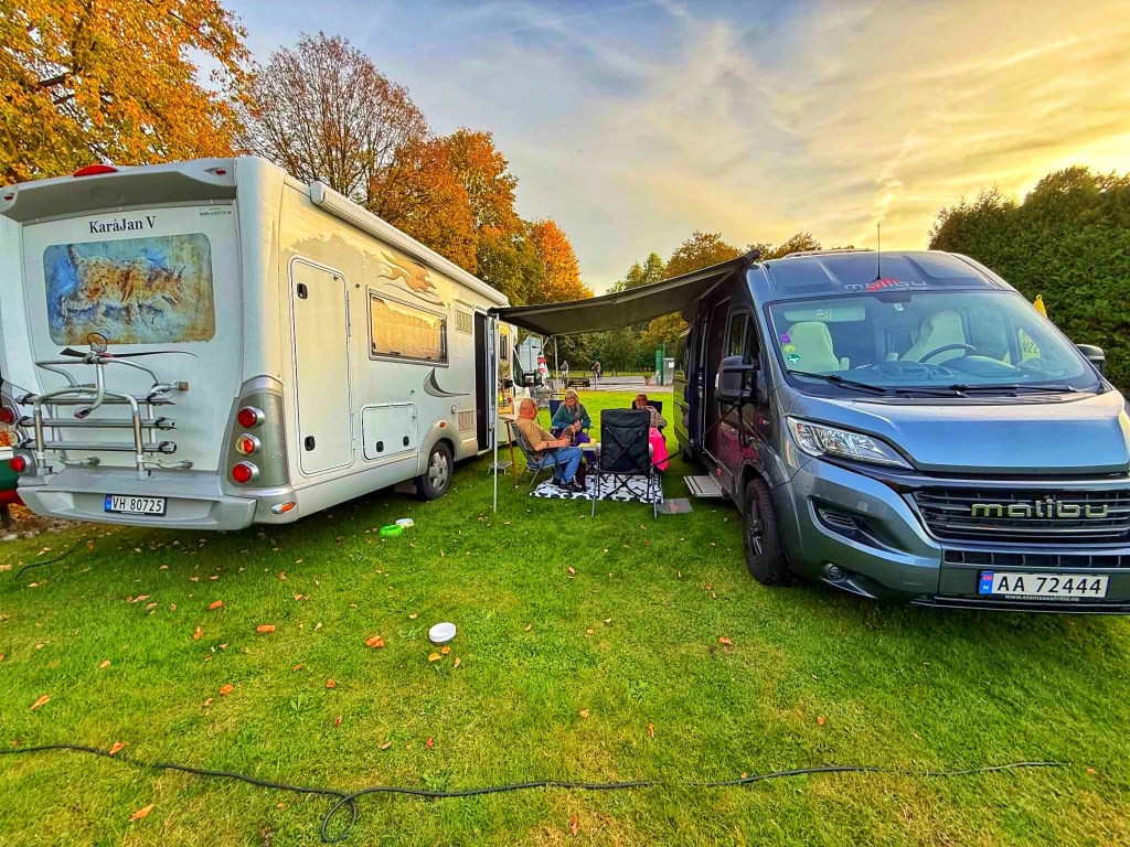 To campingvogner står på en grønn campingplass om høsten, med folk som sitter ute under markisen.