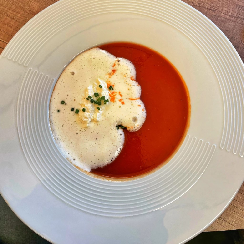 En tallerken med tomatsuppe, toppet med skum og en klatt kremost, pyntet med gressløk.