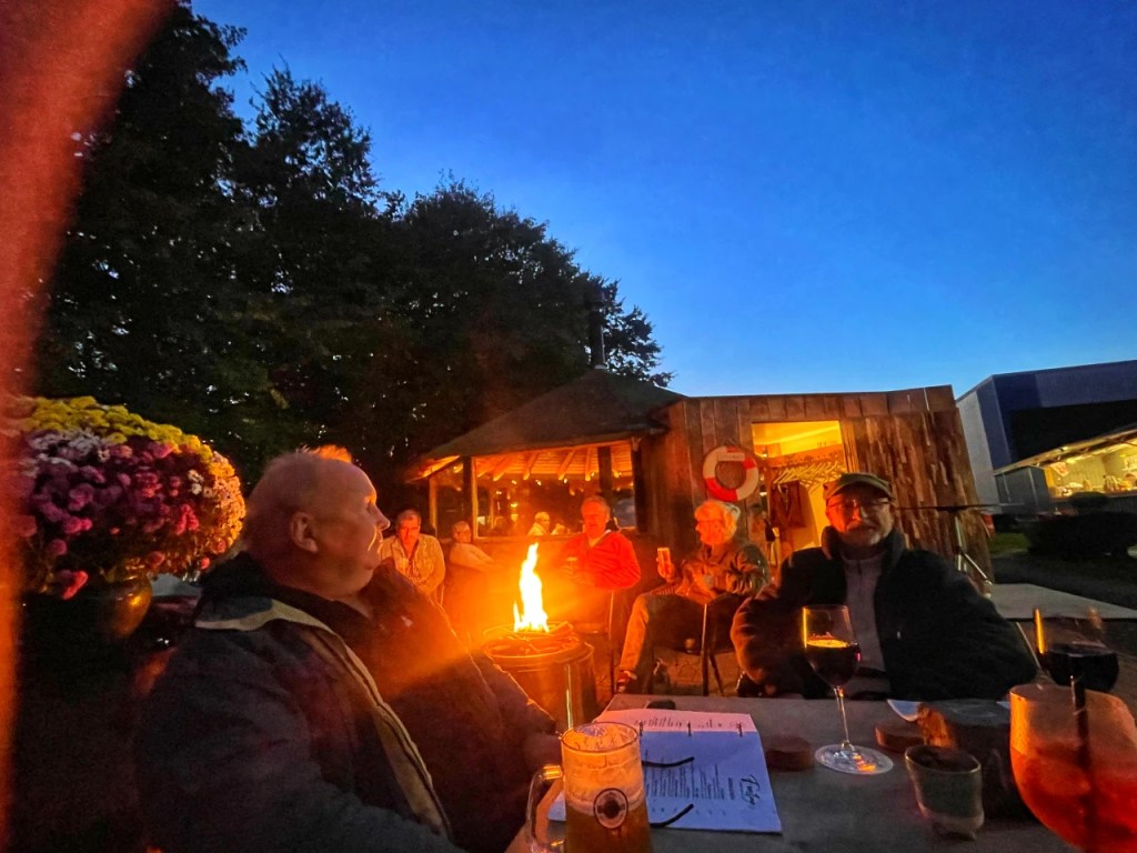 En gruppe mennesker samles rundt et bål ute på kvelden, med en mørk blå himmel i bakgrunnen. Noen holder drikker, og det er blomster i forgrunnen.