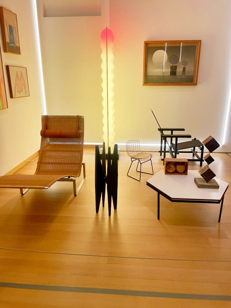 Moderne møbler i et lyst rom, inkludert en kurvestol, et lavt bord, og en stilig gulvlampe med rød topp. Det er kunstverk på veggene og et lyst tregulv.